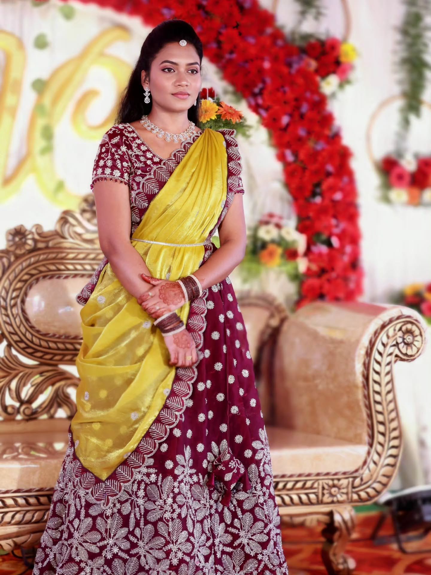 Bride in a maroon lehenga with a yellow drape at a floral reception stage