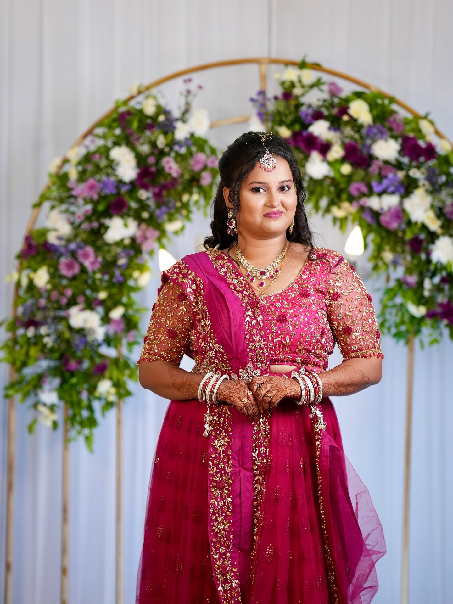 Bride in a pink lehenga beside a fresh-flower arch